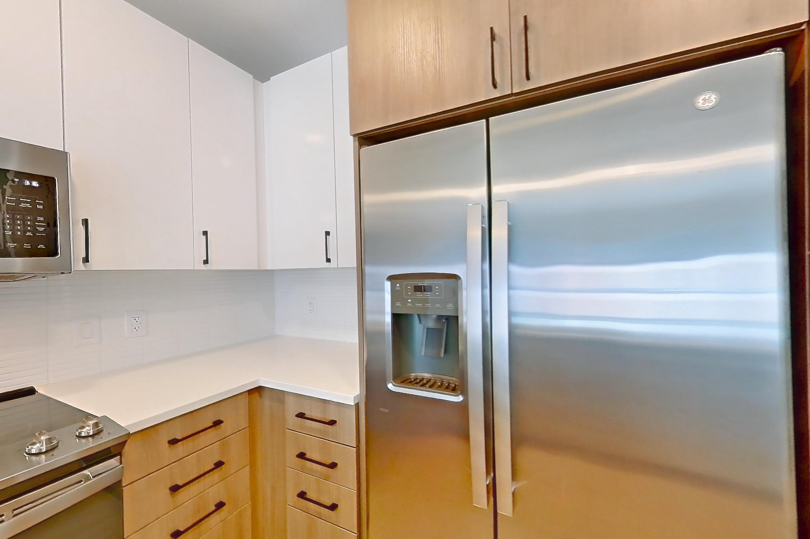 Kitchen with dishwasher, electric range, kitchen island, light countertops, light stone countertops, light wood-finish flooring, microwave, oven/range, refrigerator, stainless steel appliances, and water & ice dispenser, Apartment 0904 at The Alcott