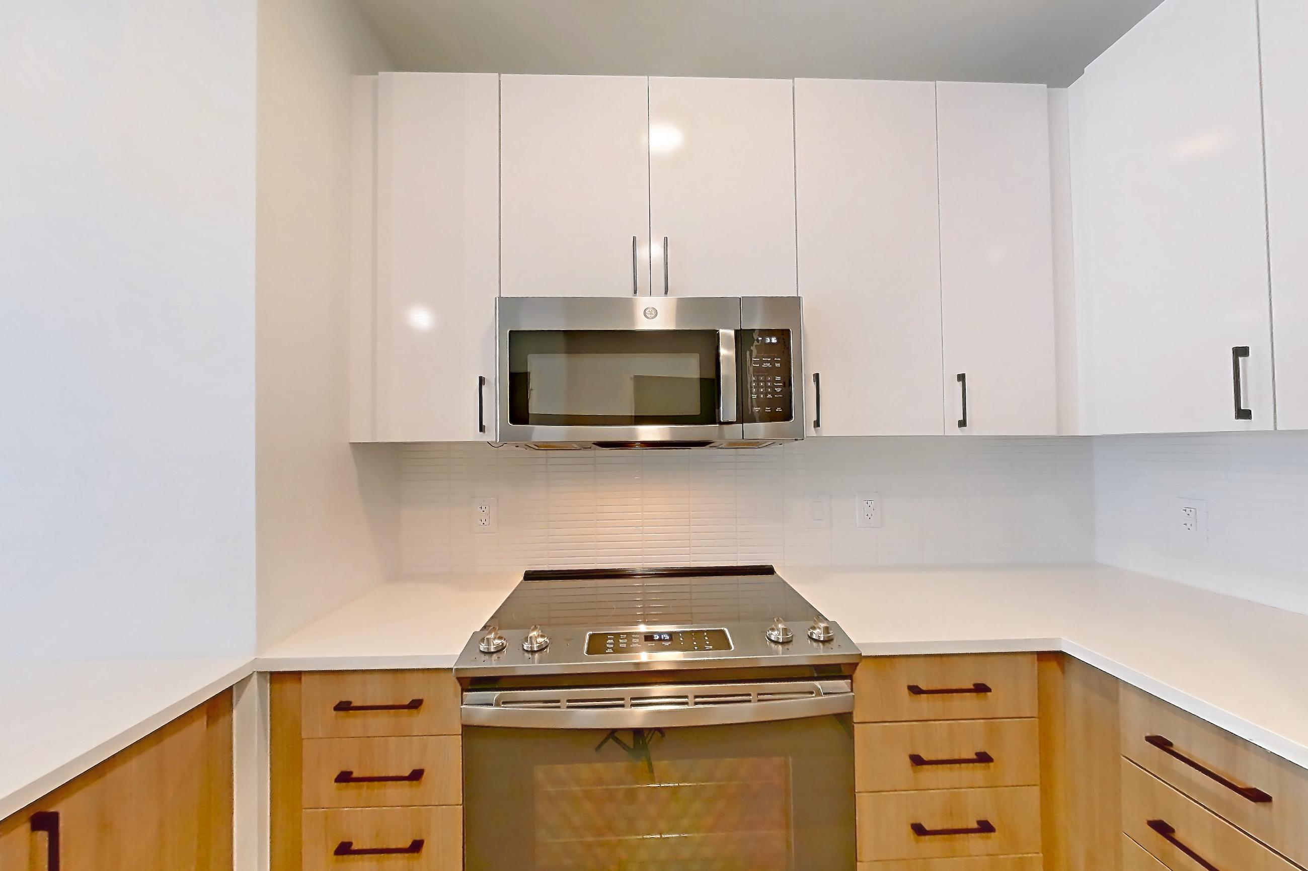 Kitchen with dishwasher, electric range, kitchen island, light countertops, light stone countertops, light wood-finish flooring, microwave, oven/range, refrigerator, stainless steel appliances, and water & ice dispenser, Apartment 0904 at The Alcott