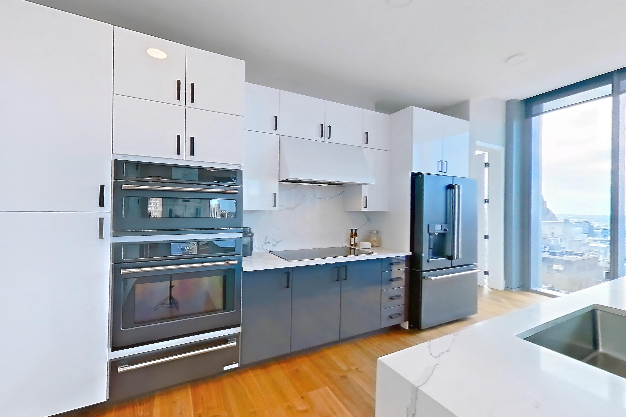 Living space with black appliances, cooktop, electric cooktop, kitchen island, light countertops, light stone countertops, light wood-finish flooring, oven/range, refrigerator, stainless steel appliances, and water & ice dispenser, Apartment 2601 at Josephine