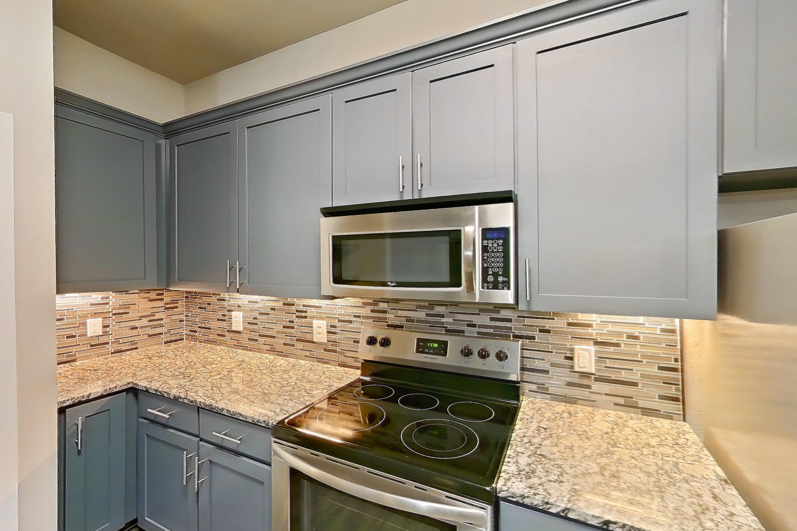 Kitchen with dark wood-finish flooring, dishwasher, electric range, kitchen island, light countertops, light stone countertops, light wood-finish flooring, microwave, oven/range, refrigerator, stainless steel appliances, and water & ice dispenser, Apartment 144 at Voyager at the Space Center