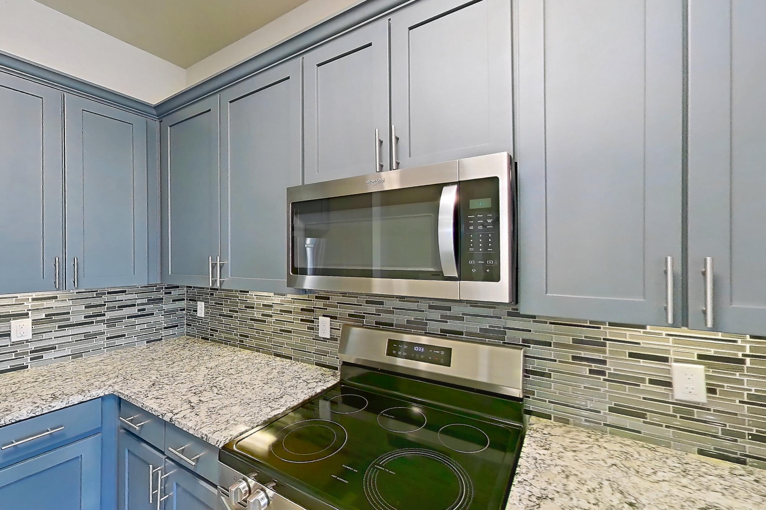 Kitchen with dark wood-finish flooring, dishwasher, electric range, kitchen island, light countertops, light stone countertops, light wood-finish flooring, microwave, oven/range, and stainless steel appliances, Apartment 410 at Voyager at the Space Center