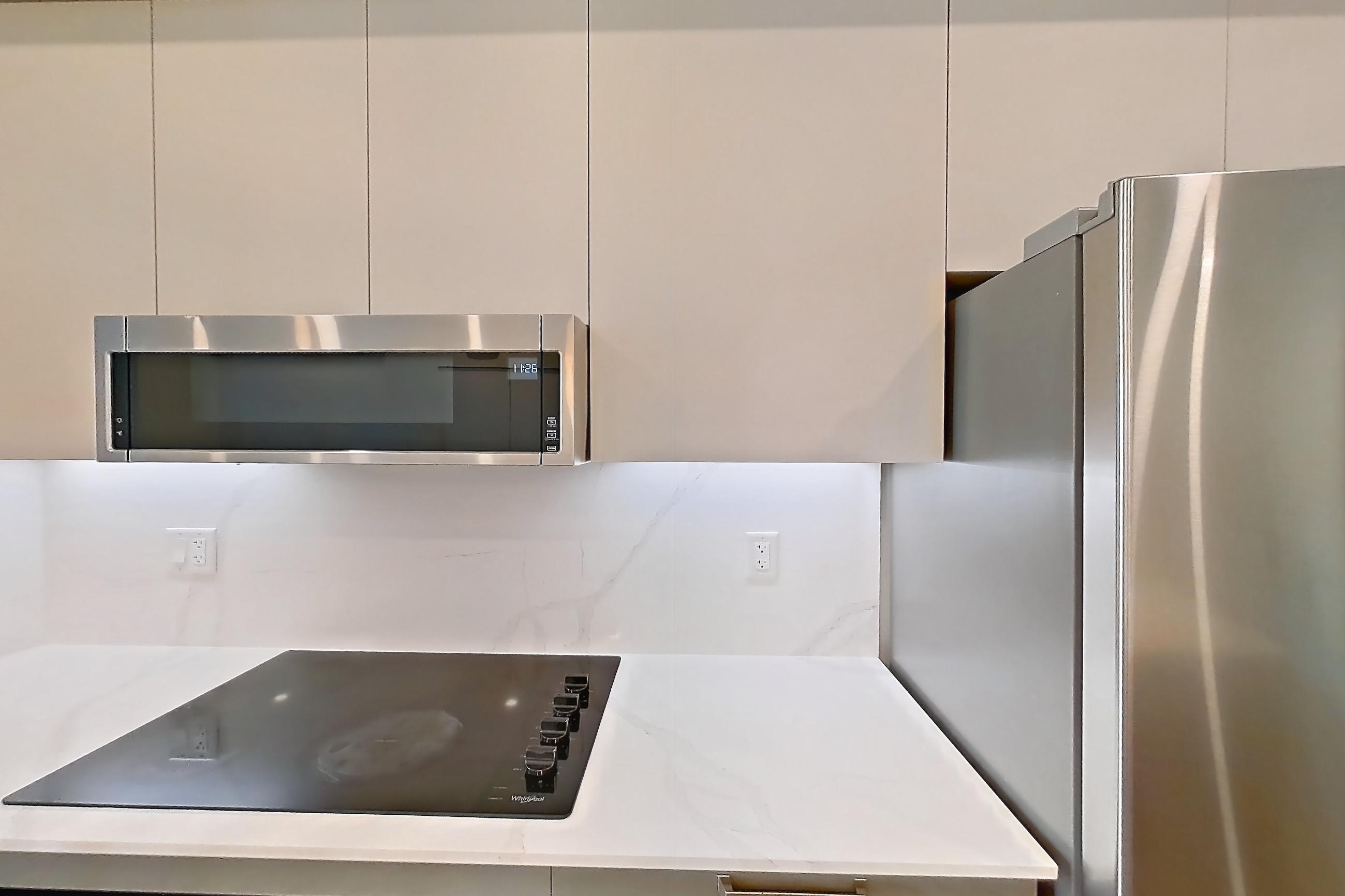 Kitchen with baseboard heating, black appliances, cooktop, dishwasher, electric cooktop, light countertops, light stone countertops, light wood-finish flooring, oven/range, refrigerator, and stainless steel appliances, Apartment T-074 at Hendry Place