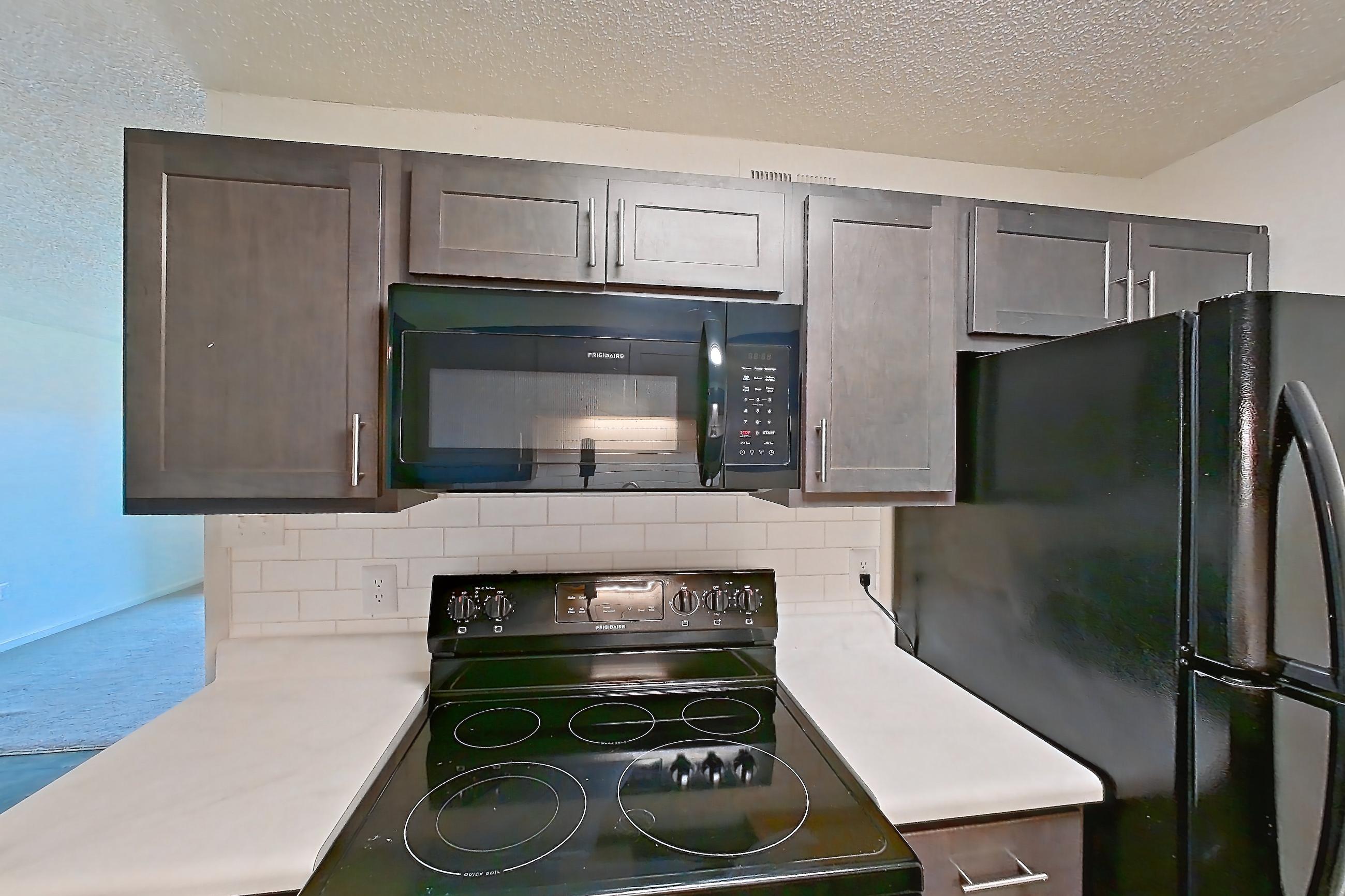 Kitchen with black appliances, dark wood-finish flooring, dishwasher, electric range, light countertops, light wood-finish flooring, microwave, oven/range, refrigerator, and stainless steel appliances, Apartment 64 at Woodwinds