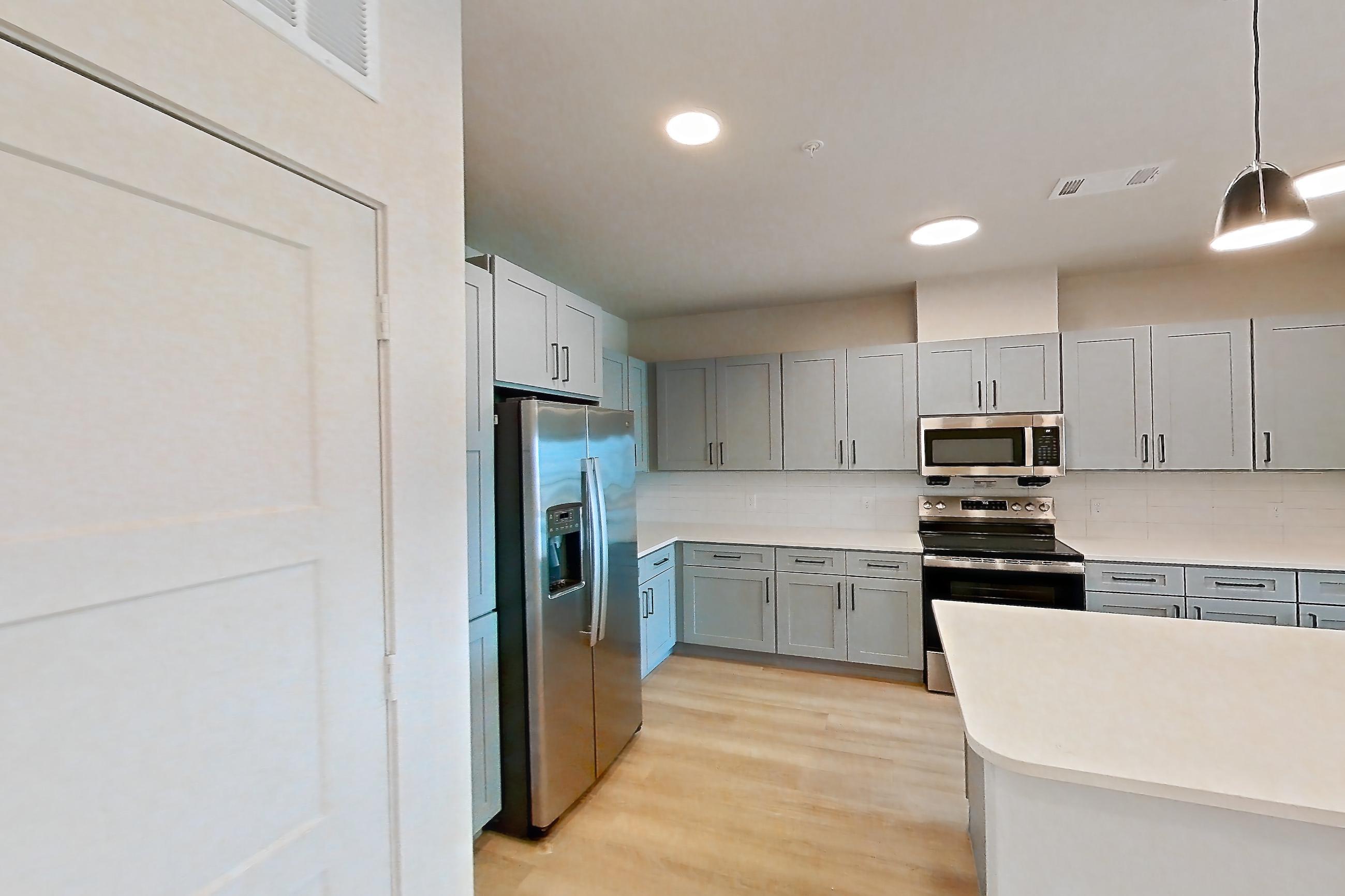 Kitchen corner with electric range, kitchen island, light countertops, light stone countertops, light wood-finish flooring, oven/range, refrigerator, stainless steel appliances, and water & ice dispenser, Apartment 3X at Residences of Siena South