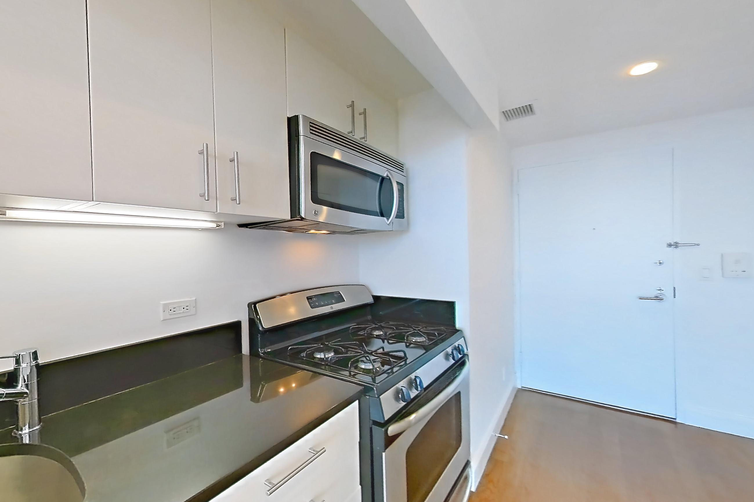 Kitchen with dark countertops, dishwasher, gas range, light countertops, light wood-finish flooring, microwave, oven/range, refrigerator, stainless steel appliances, and white appliances, Apartment 10M at The Addison