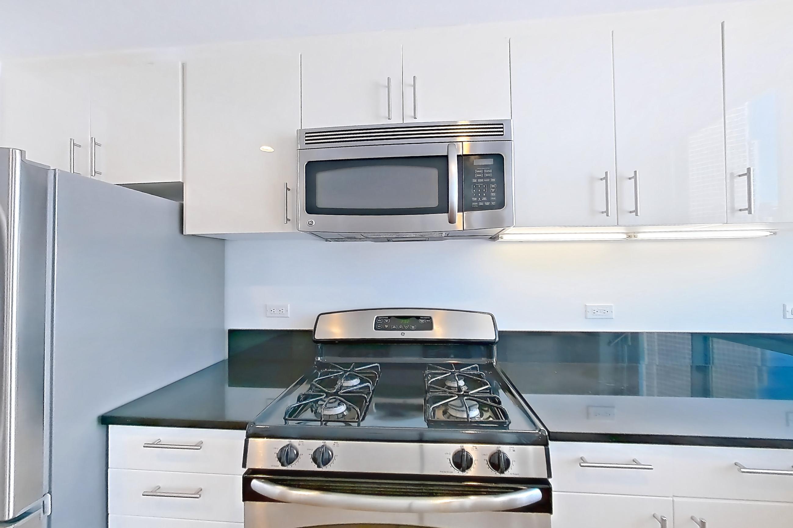Kitchen with dark countertops, dark stone countertops, dishwasher, gas range, light countertops, light wood-finish flooring, microwave, oven/range, refrigerator, stainless steel appliances, and white appliances, Apartment 18A at The Addison