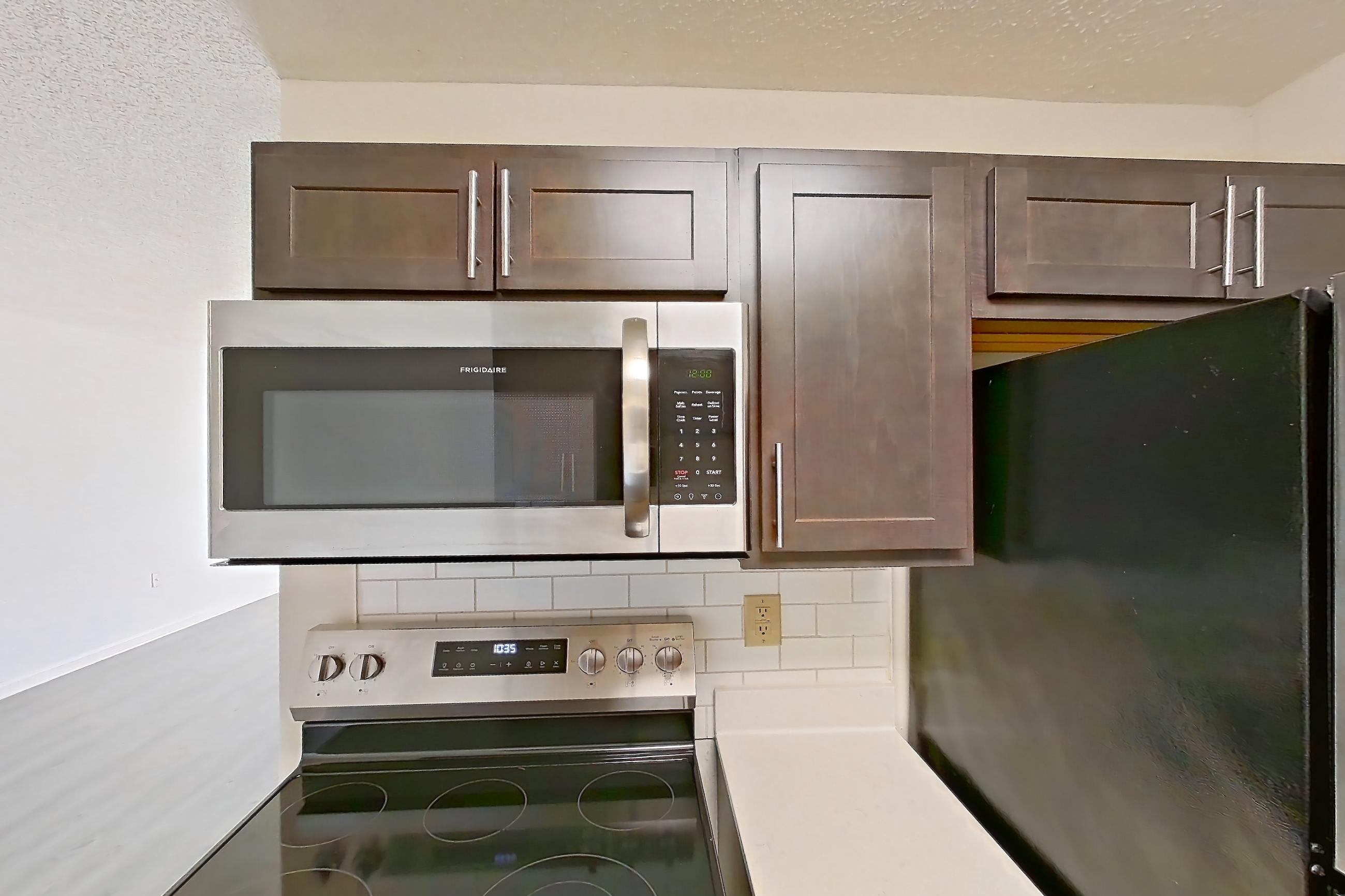 Kitchen with dark wood-finish flooring, electric range, light countertops, light stone countertops, light wood-finish flooring, microwave, oven/range, refrigerator, and stainless steel appliances, Apartment 2225-B at The Landings