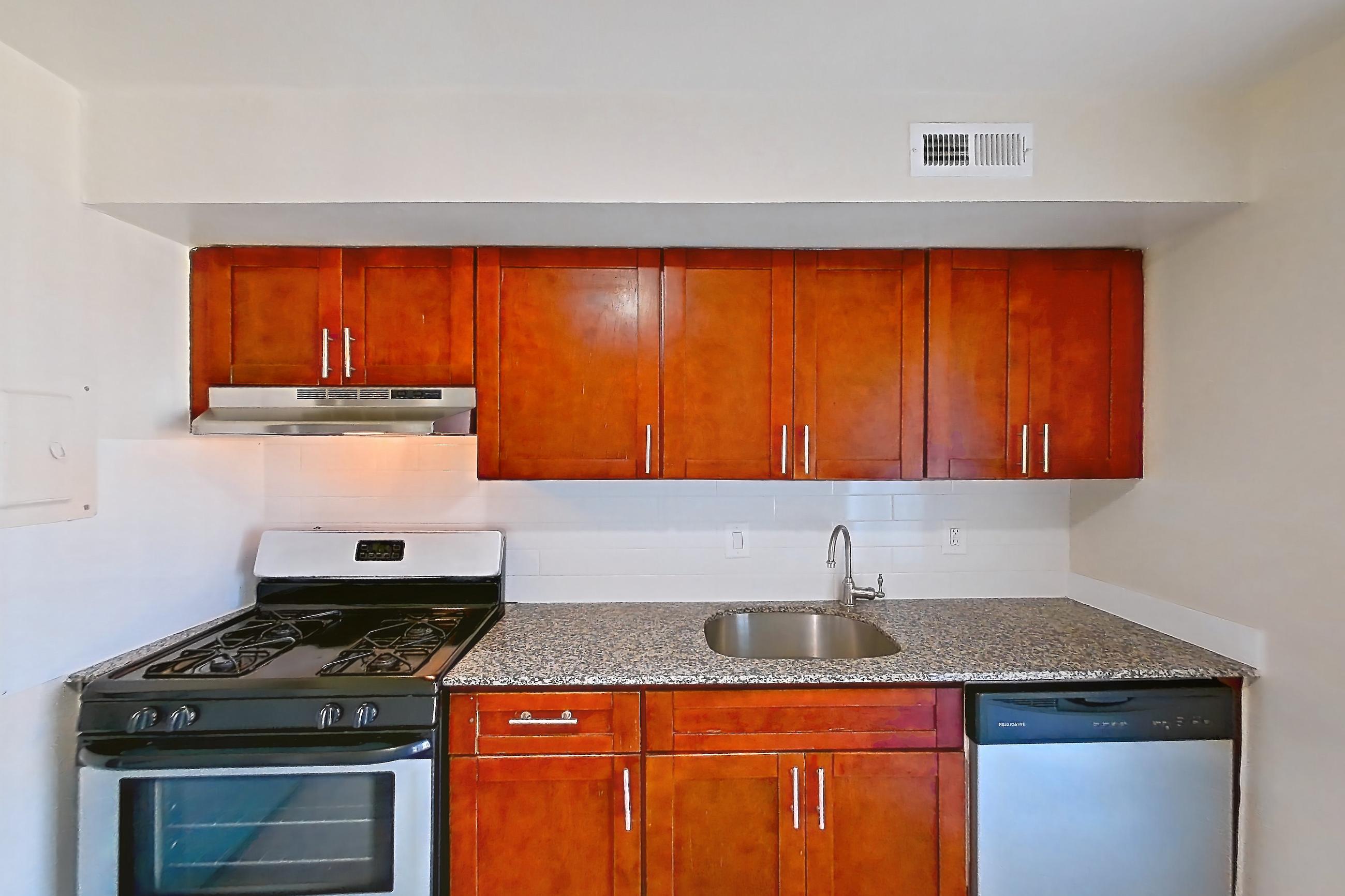 Kitchen with dark stone countertops, dishwasher, gas range, light countertops, light wood-finish flooring, oven/range, range hood, refrigerator, and stainless steel appliances, Apartment 5807CT32 at Franklin Park at Greenbelt Station