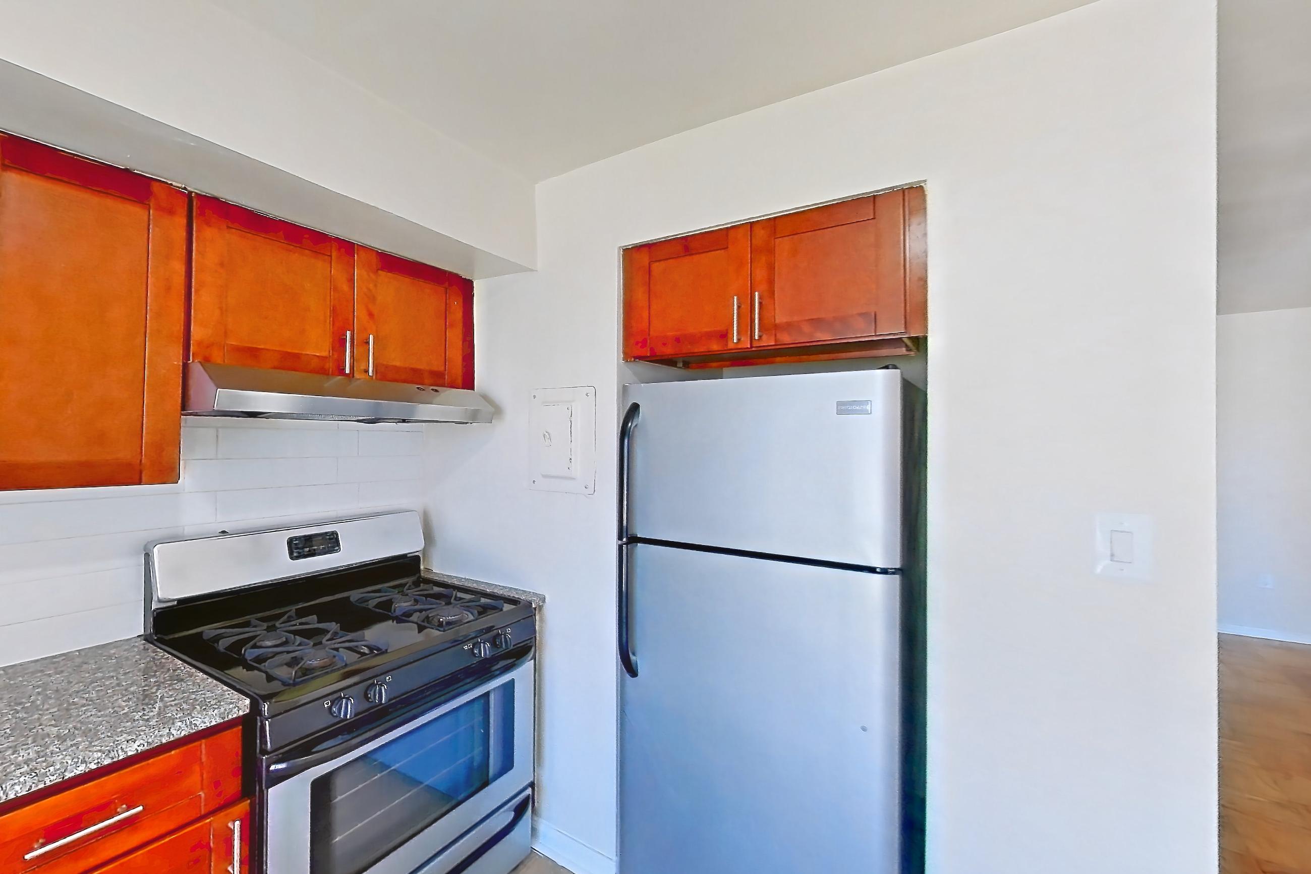 Kitchen with dark stone countertops, dishwasher, gas range, light countertops, light wood-finish flooring, oven/range, range hood, refrigerator, and stainless steel appliances, Apartment 5807CT33 at Franklin Park at Greenbelt Station