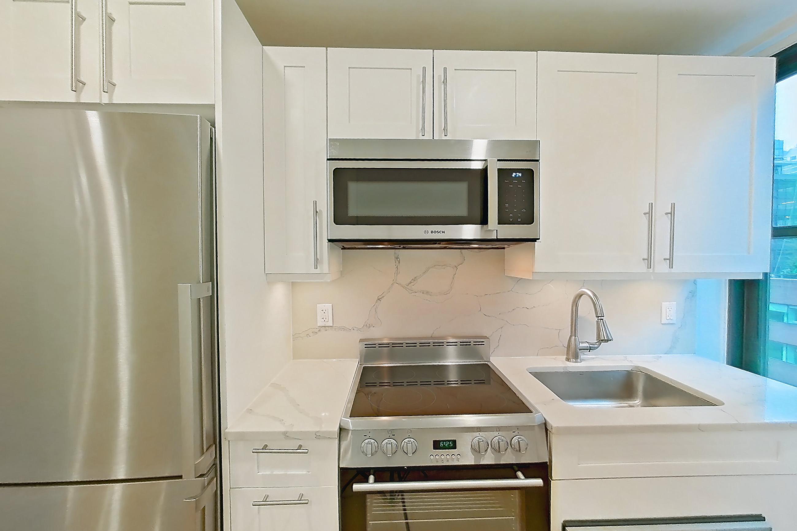 Kitchen with electric range, light countertops, light stone countertops, light wood-finish flooring, microwave, oven/range, refrigerator, and stainless steel appliances, Apartment 10K at The Buchanan