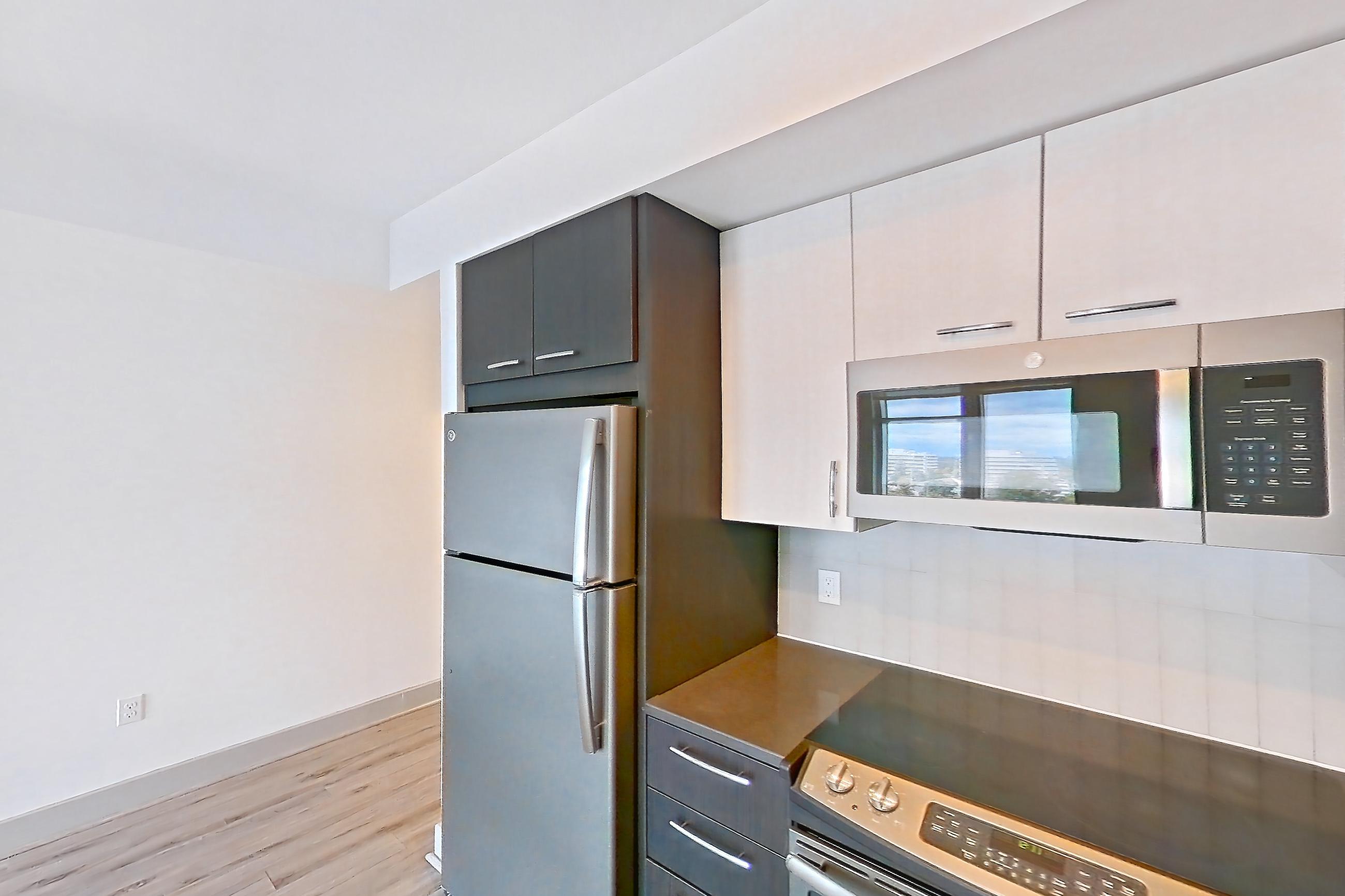 Kitchen with dark countertops, dark stone countertops, dishwasher, electric range, light countertops, light wood-finish flooring, microwave, oven/range, refrigerator, stainless steel appliances, and white appliances, Apartment 906 at Adaire