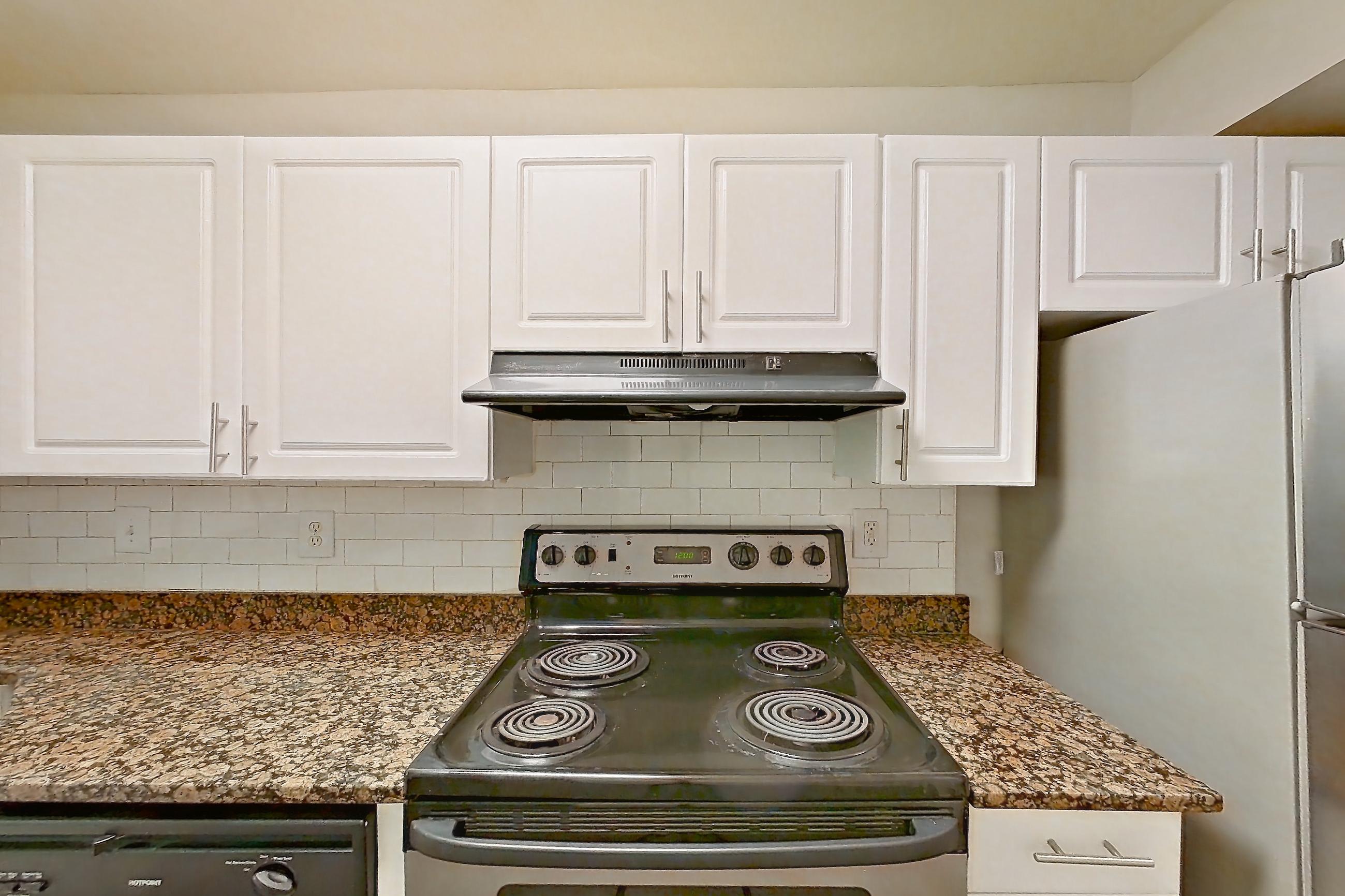 Kitchen with dark stone countertops, dishwasher, electric range, light countertops, light stone countertops, light wood-finish flooring, oven/range, range hood, refrigerator, stainless steel appliances, and white appliances, Apartment 3558W at The Ashford